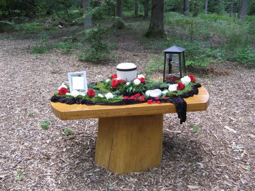 Urne auf einem Holzaltar bei einer Waldbestattung zwischen Bäumen als Ort für Abschied und persönliche Erinnerung.